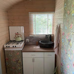 Compact kitchenette with gas hob, black basin sink, wooden countertops, and floral wallpaper accents in a rustic hut interior.