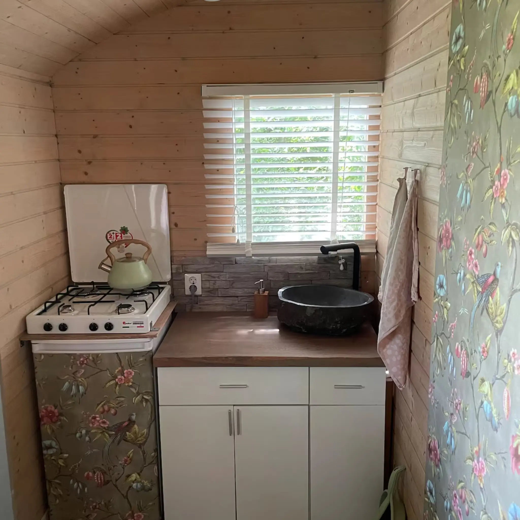 Compact kitchenette with gas hob, black basin sink, wooden countertops, and floral wallpaper accents in a rustic hut interior.