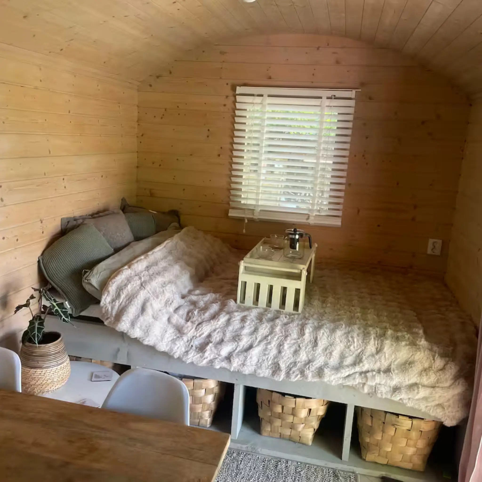 Cosy sleeping area with plush bedding, wooden interior walls, woven storage baskets, and natural light through shuttered windows.
