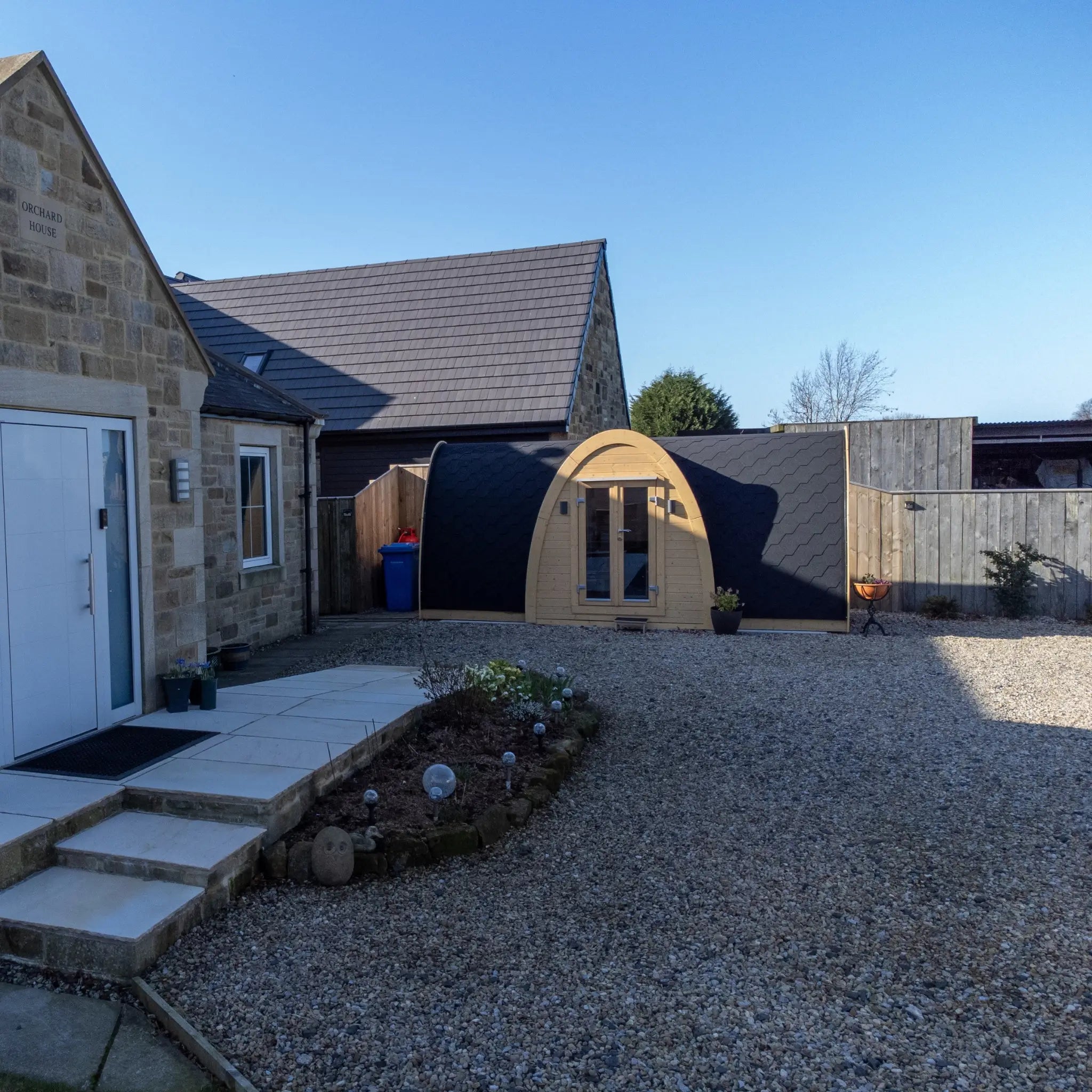 Wide front view of Hobbit garden office pod in landscaped garden setting with gravel courtyard and contemporary home backdrop