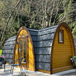 Exterior side view of Hobbit pod luxury 2 person glamping cabin with curved roof, timber cladding and forest backdrop