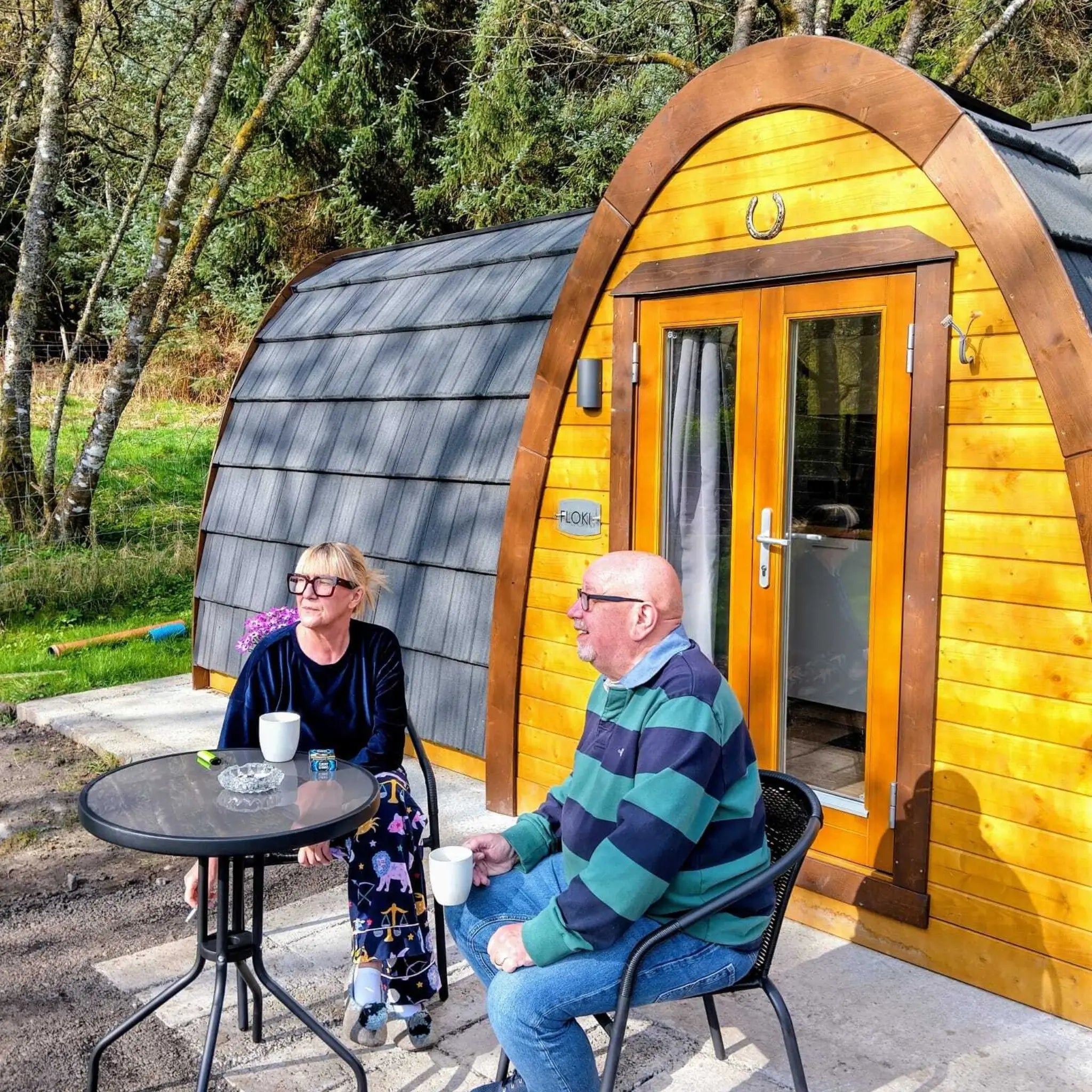 Two people enjoying coffee outside Hobbit pod glamping cabin with arched wooden entrance and outdoor seating area