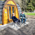 Couple relaxing outside luxury Hobbit glamping pod with patio seating, curved timber design and countryside setting