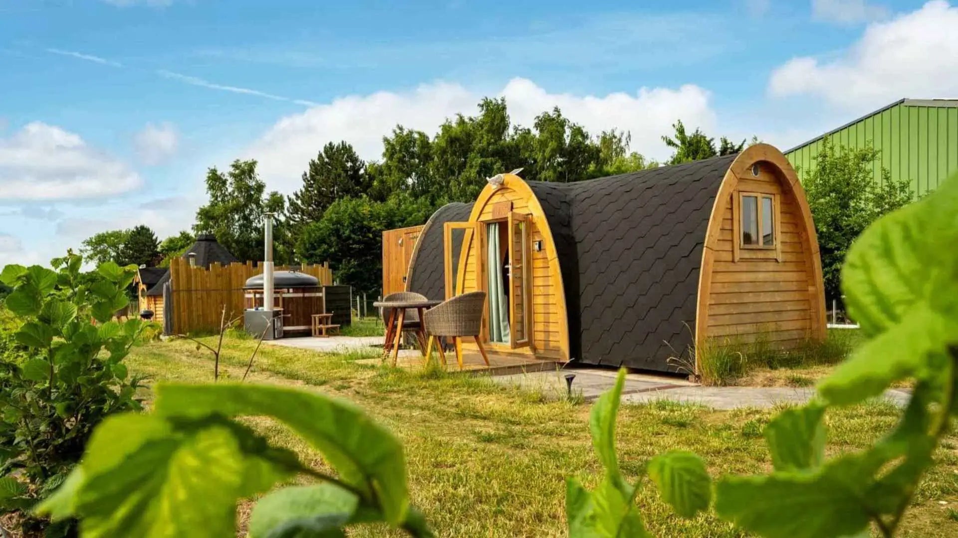 A timber-clad luxury glamping pod with curved black shingle roof, outdoor seating, and a private hot tub, set in a landscaped rural site on a sunny day.